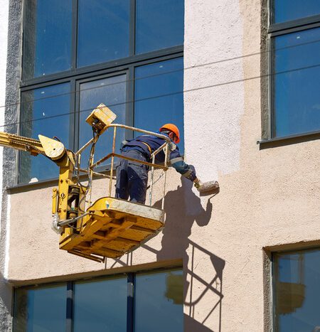 Commercial painting worker painting the wall with brush roller in lifting bucket. Painter working on crane platform, paint building wall. Men at work in hardhat paint facade of building at height in lifting cradle.